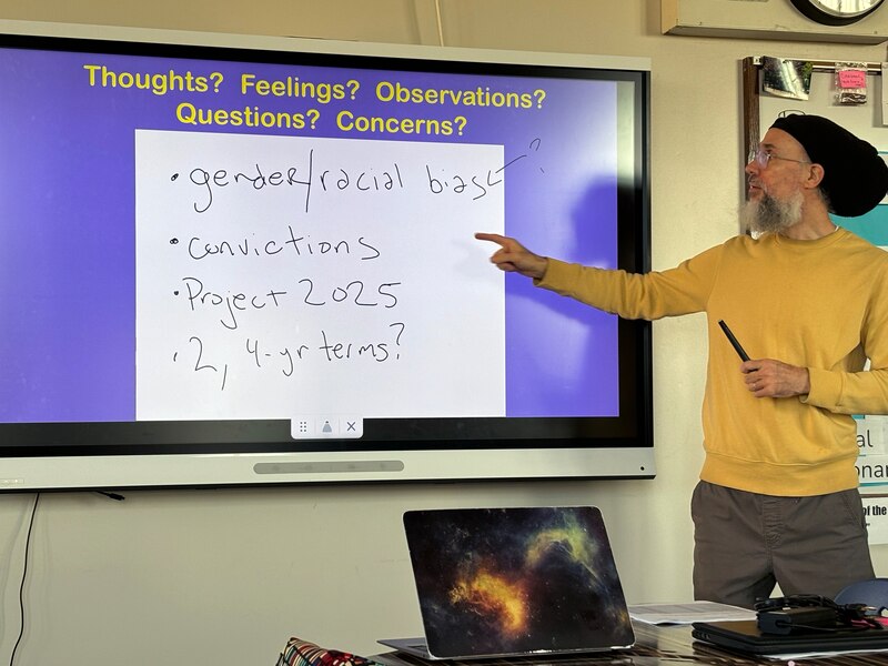 A teacher stands at the front of a class by a projected screen on a wall.