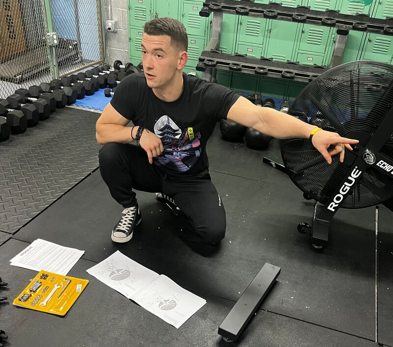 Photo of a man in a black shirt, sitting in a workout room. There are weights in the background.